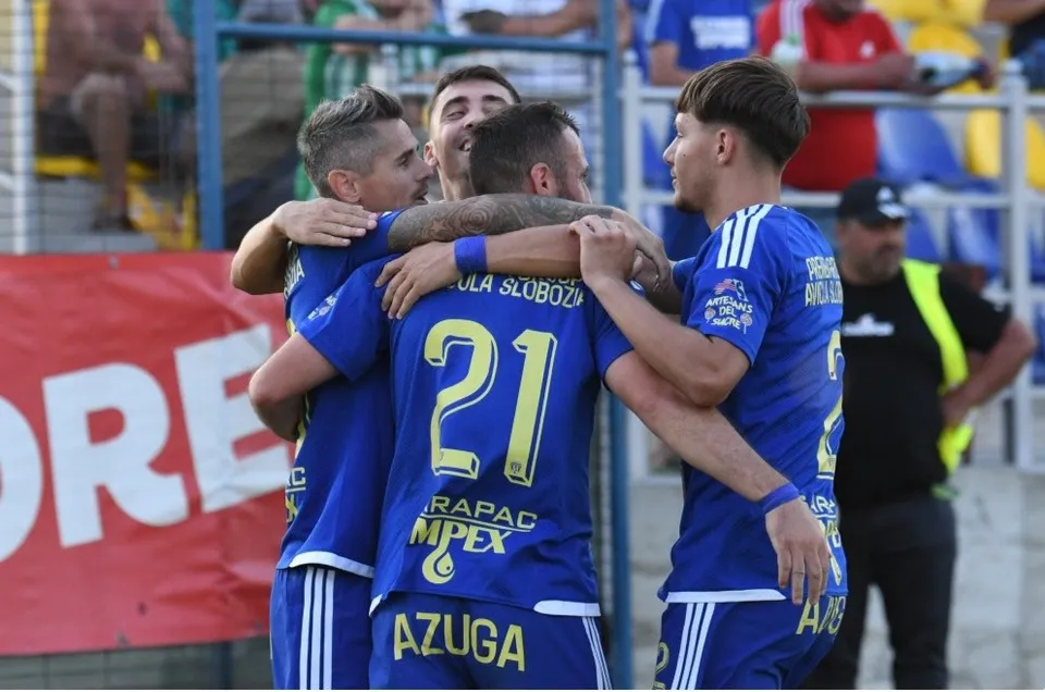 Patru jucători de fotbal în echipamente albastre se îmbrățișează pe un teren de sport.