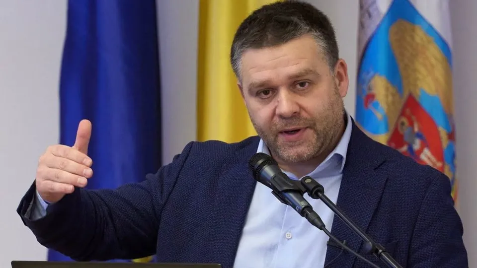 Un bărbat cu sacou închis și cămașă deschisă vorbește la un microfon în fața unor steaguri colorate.
