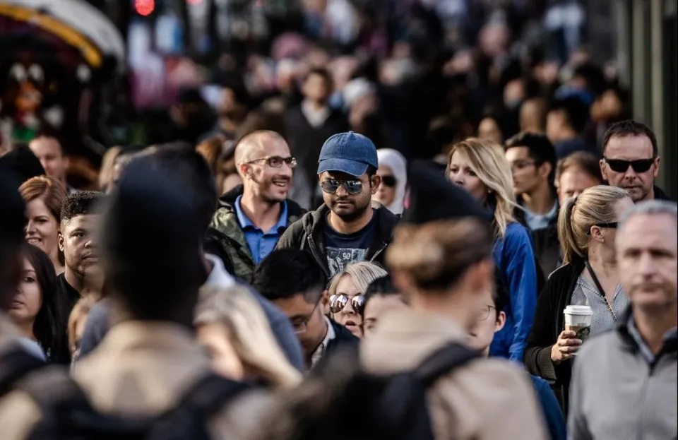 O mulțime de oameni merge pe o stradă aglomerată, printre care un bărbat cu ochelari de soare și o șapcă albastră este vizibil în centru.