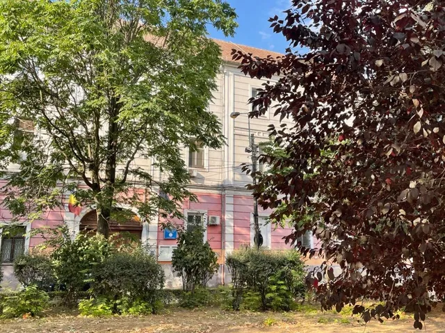 O clădire cu fațadă roz și albă este parțial acoperită de un copac verde mare și un arbust cu frunze purpurii în prim-plan.