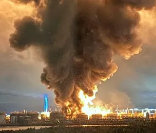 O coloană densă de fum negru și cenușiu se ridică deasupra unor rezervoare industriale de lângă un flăcările mari de foc.
