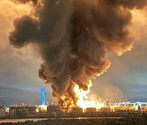 O coloană densă de fum negru și cenușiu se ridică deasupra unor rezervoare industriale de lângă un flăcările mari de foc.