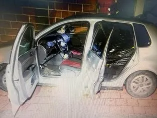 O mașină de culoare deschisă cu ambele portiere din partea stângă deschise este parcată pe un trotuar lângă un perete de cărămidă.