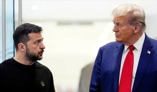 Un bărbat cu barbă poartă un tricou negru și privește spre un bărbat cu părul blond, costum albastru și cravată roșie.