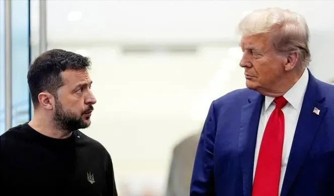 Un bărbat cu barbă poartă un tricou negru și privește spre un bărbat cu părul blond, costum albastru și cravată roșie.