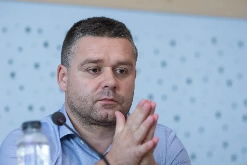 Un bărbat cu părul scurt și cămașă albastră stă la o masă cu mâinile împreunate sub bărbie în fața unui fundal albastru cu puncte.