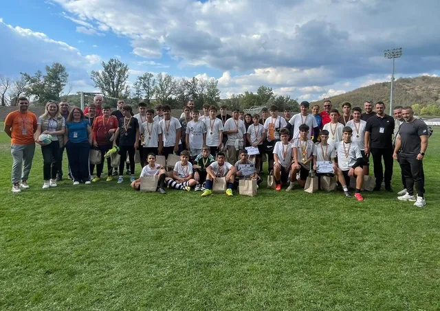 Un grup mare de copii și adulți stau în rând pe un teren de fotbal verde sub un cer înconjurat de nori.