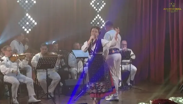 Un grup de muzicieni în haine albe și o femeie în costum popular cântă și dansează pe o scenă iluminată.