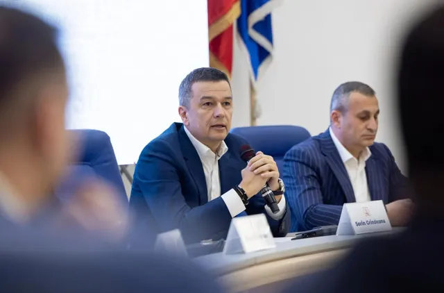 Un bărbat într-un costum albastru stă la o masă și vorbește, în timp ce un alt bărbat în costum în dungi îl ascultă lângă el.