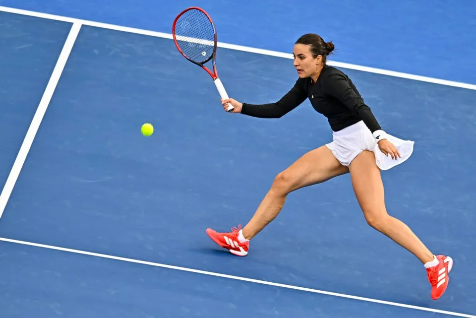 O femeie cu părul negru strâns într-o coadă de cal lovește o minge galbenă cu o rachetă pe un teren de tenis albastru.