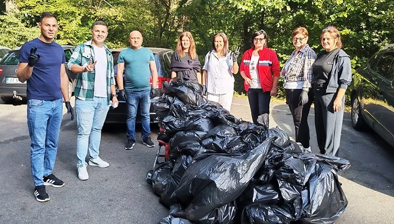 Un grup de oameni stă lângă un munte de saci negri de gunoi pe un drum asfaltat, în fața unor copaci verzi.