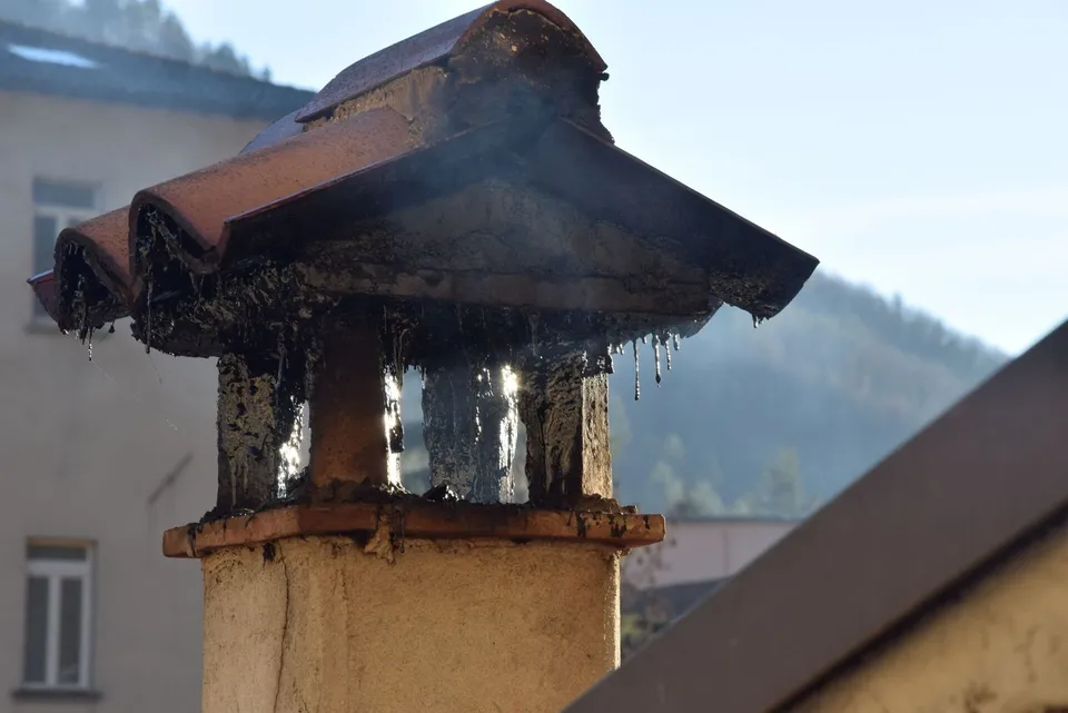 Un coș de fum metalic cu acoperiș este montat pe un suport de beton sub un cer luminos.