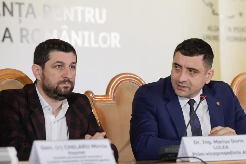 Un bărbat cu sacou albastru și cravată neagră vorbește la o masă lângă un bărbat cu sacou în carouri și cămașă albă.