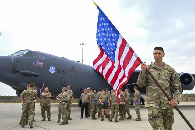 Un soldat în uniformă de camuflaj ține un steag american în fața unui avion militar gri pe o pistă de aterizare cu alți militari în fundal.