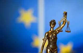 O statuie de bronz a unei femei ținând o balanță este poziționată în fața unui fundal albastru cu stele galbene difuze.