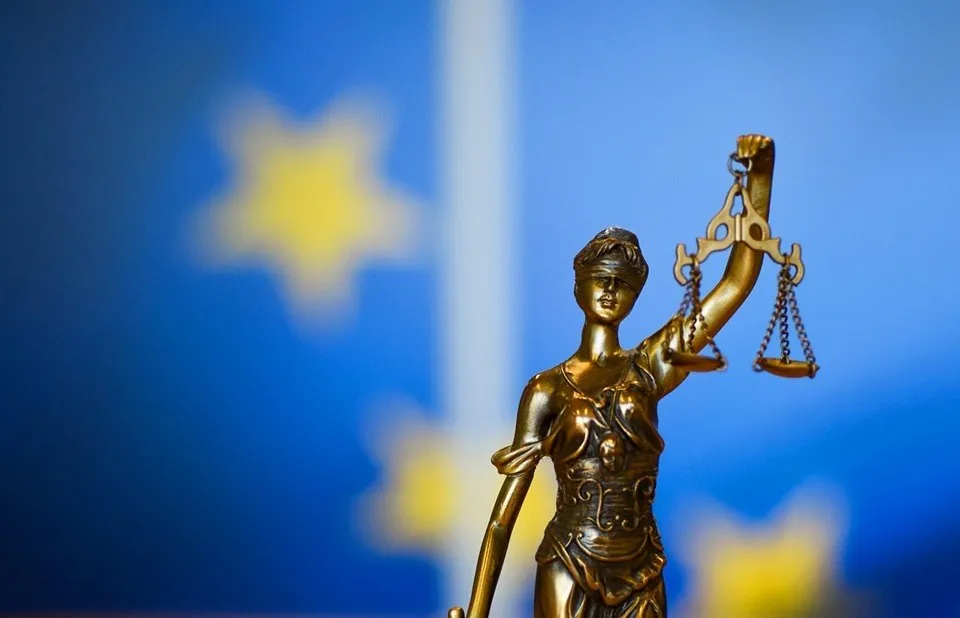 O statuie de bronz a unei femei ținând o balanță este poziționată în fața unui fundal albastru cu stele galbene difuze.