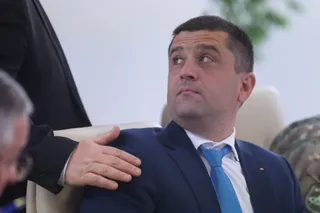 Un bărbat cu păr scurt, îmbrăcat în costum albastru și cravată albastră, stă așezat într-un fotoliu cu o mână a unei alte persoane pe umărul său.