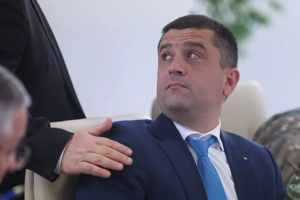 Un bărbat cu păr scurt, îmbrăcat în costum albastru și cravată albastră, stă așezat într-un fotoliu cu o mână a unei alte persoane pe umărul său.