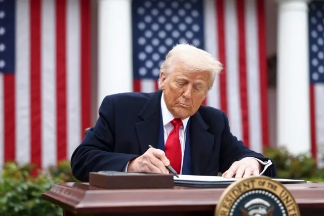 Bărbat cu păr blond, costum închis și cravată roșie stă la un birou, semnând un document, cu steaguri americane și coloane albe în fundal.