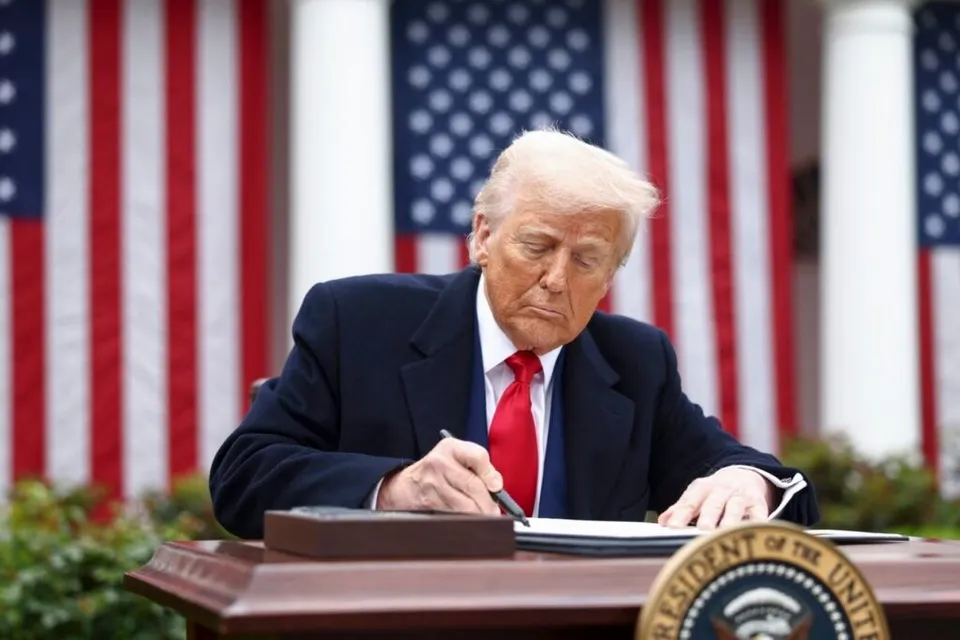 Bărbat cu păr blond, costum închis și cravată roșie stă la un birou, semnând un document, cu steaguri americane și coloane albe în fundal.