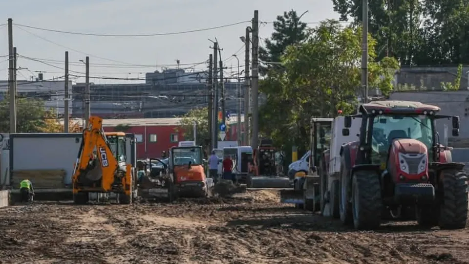 Mai multe utilaje de construcții, inclusiv un excavator portocaliu și un tractor roșu, sunt poziționate pe un teren de pământ întins.