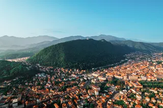 O vedere aeriană a unui oraș cu case cu acoperișuri roșii, situat la baza unor dealuri verzi sub un cer senin.