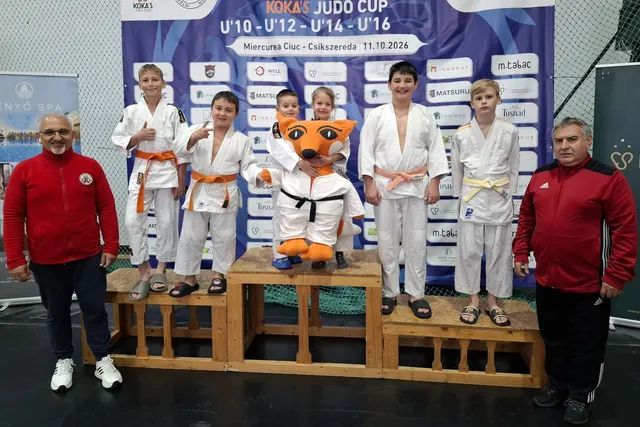 Șapte copii îmbrăcați în uniforme albe de judo stau pe podium lângă doi bărbați în haine sportive roșii și negre.