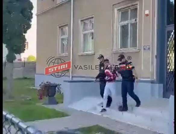 Trei persoane merg pe o cărare lângă o clădire cu ferestre înalte, în timp ce un logo de știri este suprapus pe imagine.
