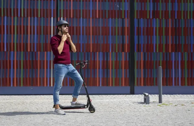 Un bărbat cu cască și ochelari de soare stă pe o trotinetă electrică în fața unui perete colorat cu dungi verticale.