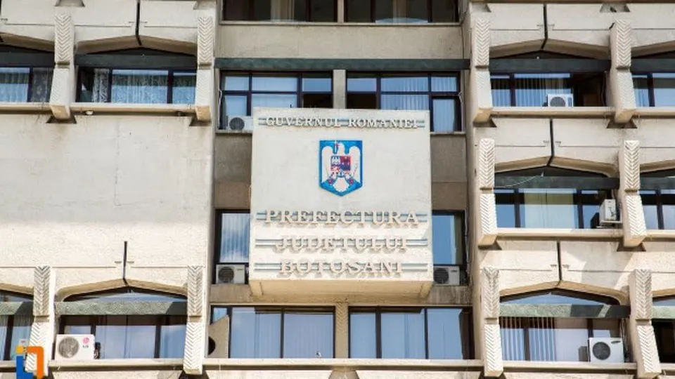 O clădire oficială cu un panou mare pe care scrie Prefectura Județului Botoșani, având un stema albastru și roșu în centru.