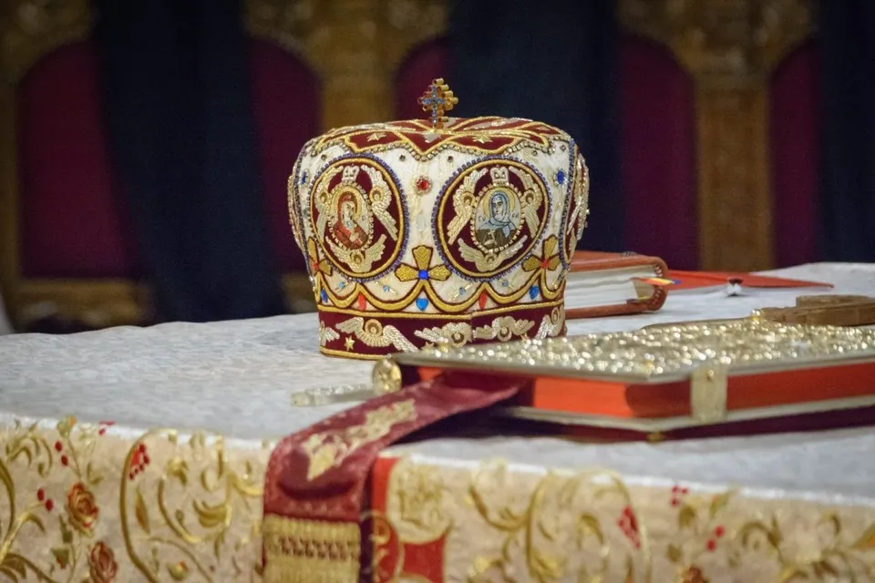 O mitră religioasă decorată cu iconițe și pietre colorate stă pe o masă acoperită cu un drapu de culoare deschisă lângă o carte roșie.