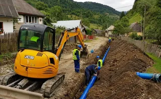 O excavatoare galbenă de mici dimensiuni lucrează lângă un șanț în care sunt așezate țevi de culoare albastră, în prezența unor lucrători.