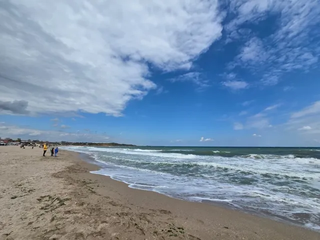 O plajă cu nisip se întinde sub un cer albastru cu nori, având valuri albe care se sparg spre mal.