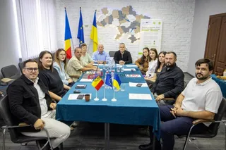 Un grup de oameni stă așezat în jurul unei mese acoperite cu un textil albastru într-o încăpere cu un hartă pe perete și steaguri.