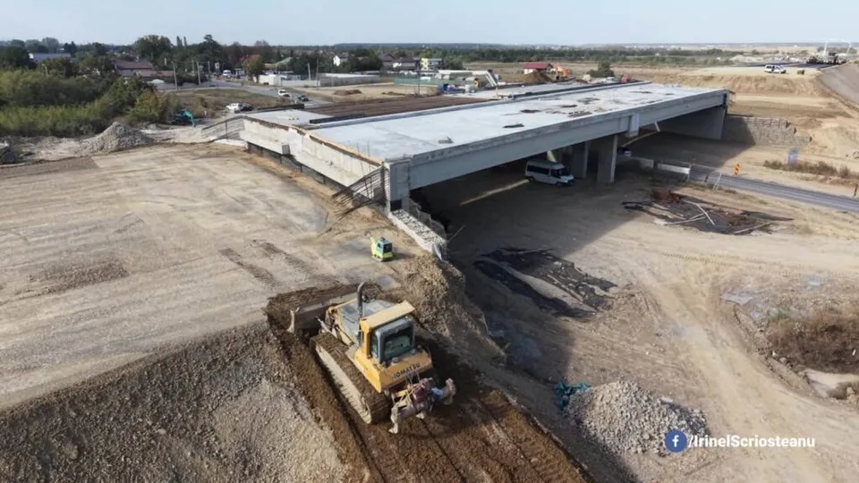 O utilaj de construcții galben lucrează pe un teren de pământ lângă o structură de viaduct de beton în construcție.