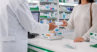Un farmacist într-un halat alb întinde o cutie mică unui client aflat în fața unui raft cu diverse produse medicamentoase.