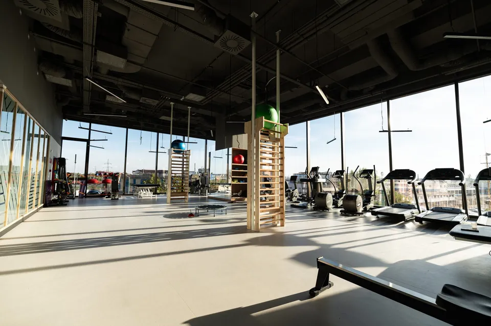 O sală de fitness cu ferestre mari, echipamente de antrenament și structuri din lemn dispuse pe un podea deschisă sub un tavan industrial întunecat.