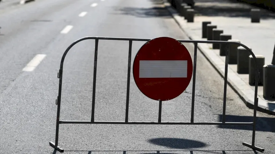 Un gard metalic cu un semn circular roșu și o bară albă este poziționat pe marginea unei străzi asfaltate.