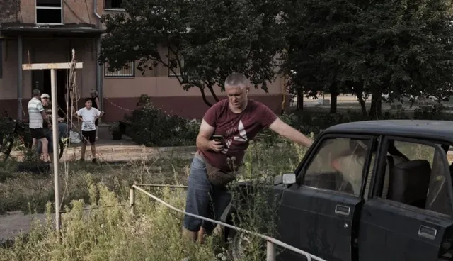 Un bărbat cu tricou roșu și pantaloni albaștri stă lângă o mașină deschisă într-o zonă cu vegetație înaltă.