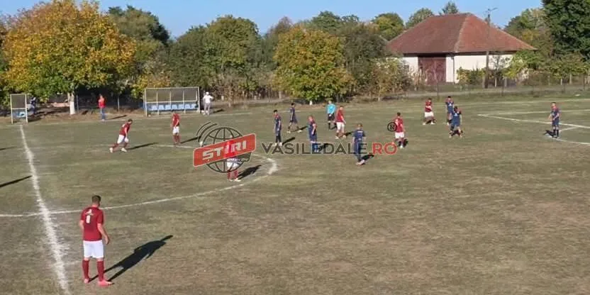 Un grup de jucători de fotbal în echipamente roșii și albastre se află pe un teren de sport cu iarbă uscată sub un cer senin.