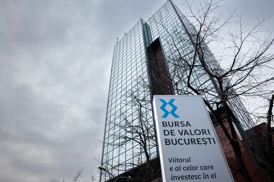O clădire înaltă cu fațadă de sticlă este vizibilă sub un cer înnorat, lângă un panou informativ alb cu text albastru și negru.