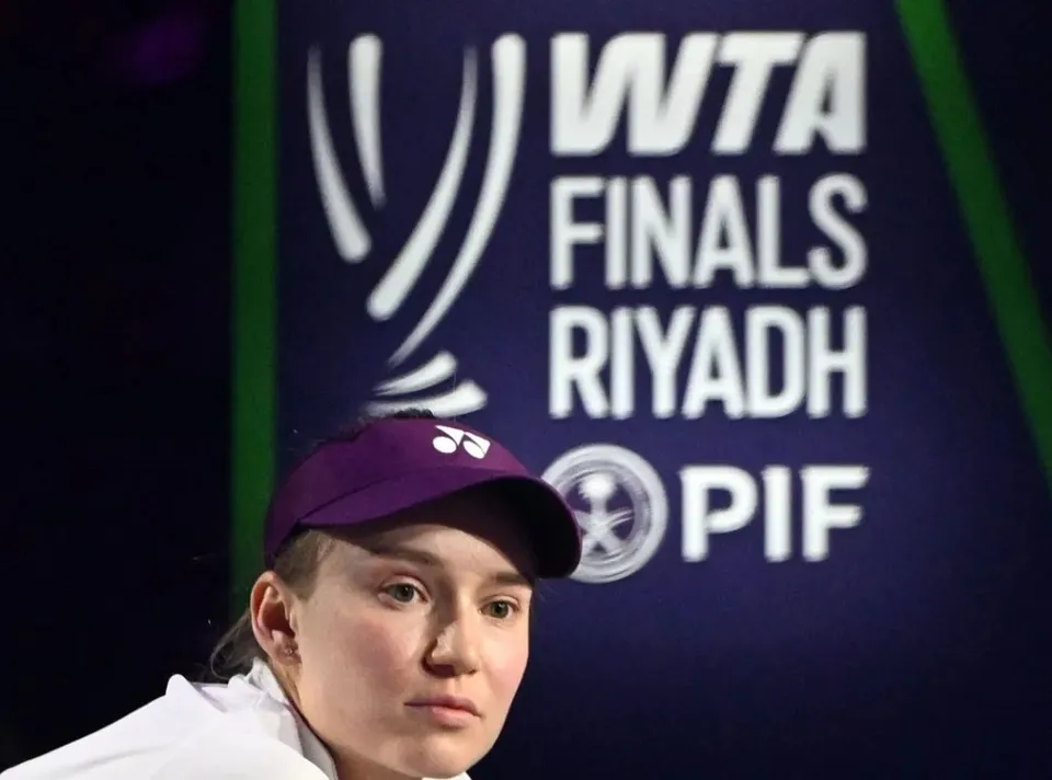 O femeie cu șapcă mov și tricou alb privește în lateral în fața unui panou cu textul WTA Finals Riyadh.