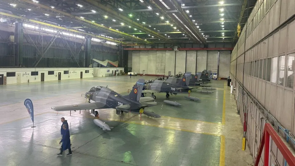 Mai multe avioane de training de culoare închisă sunt parcate într-un hangar mare cu podea verde și un om merge pe lângă ele.