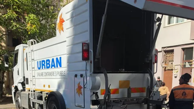Un camion alb cu inscripția URBAN și un simbol de frunză este parcat pe o stradă lângă două persoane.