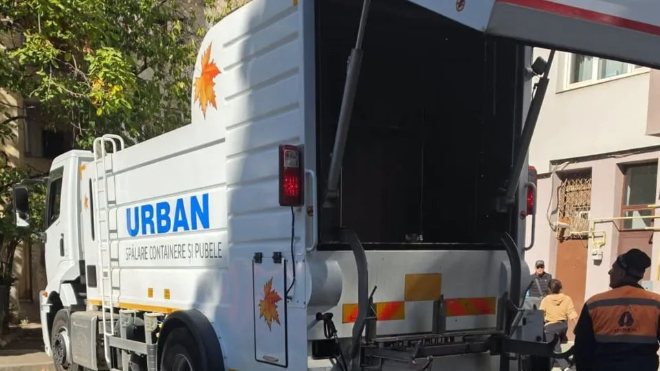 Un camion alb cu inscripția URBAN și un simbol de frunză este parcat pe o stradă lângă două persoane.