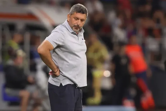 Un bărbat cu părul scurt și barbă căruntă poartă un tricou polo gri și pantaloni închiși, stând cu mâinile pe șolduri într-un stadion.
