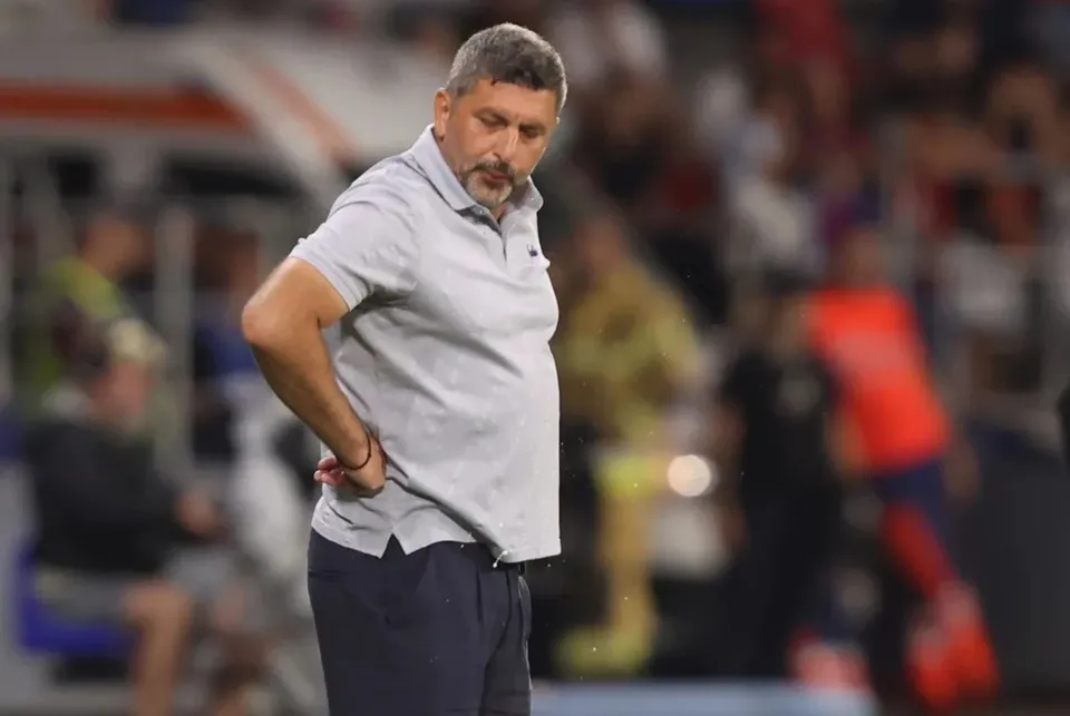 Un bărbat cu părul scurt și barbă căruntă poartă un tricou polo gri și pantaloni închiși, stând cu mâinile pe șolduri într-un stadion.
