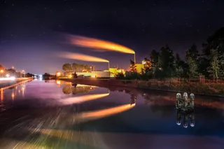 Două coloane de fum portocaliu ies din hornuri industriale și se reflectă în apa de lângă un canal sub un cer nocturn.