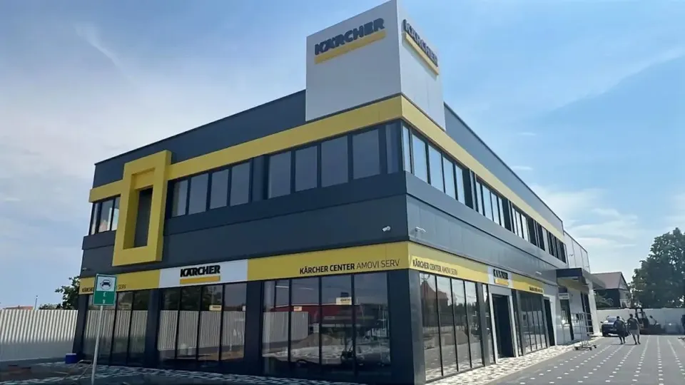 O clădire modernă cu două etaje prezintă fațade gri și galben, având ferestre largi și un panou publicitar cu textul Kärcher pe partea superioară.