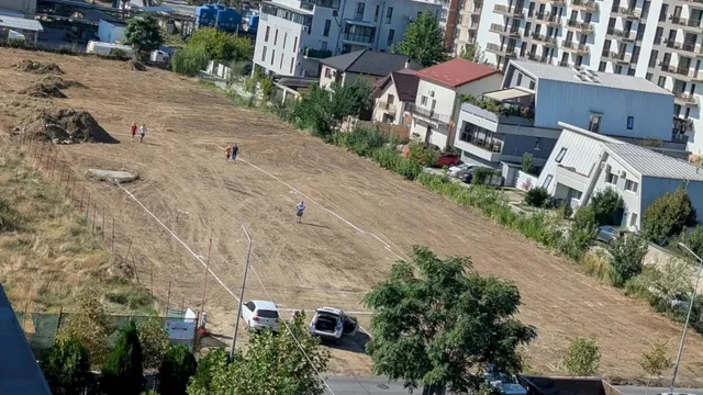 Un teren gol cu iarbă uscată este delimitat de un gard, având câteva persoane și mașini parcate în apropiere de clădiri rezidențiale.